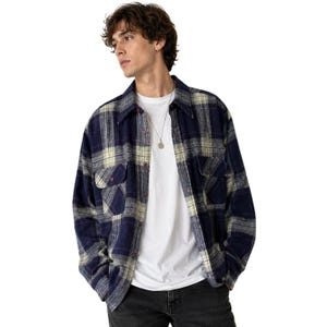Könnte beinhalten: Ein marineblaues und cremefarbenes Flanellhemd mit zwei Brusttaschen. Das Hemd ist geknöpft und wird über einem weißen T-Shirt getragen. Das Model trägt eine schwarze Jeans und eine silberne Halskette.