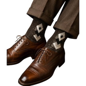 May include: A pair of brown leather dress shoes with matching laces. The shoes are paired with dark gray socks featuring a white diamond pattern. The socks are visible beneath brown trousers with cuffed hems.