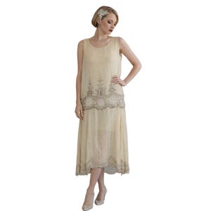 May include: A cream-coloured, sleeveless flapper dress with intricate beaded embellishments. The dress features a layered design with a detailed border at the waist and hem. The model is wearing a matching headpiece and heels, showcasing the vintage style.