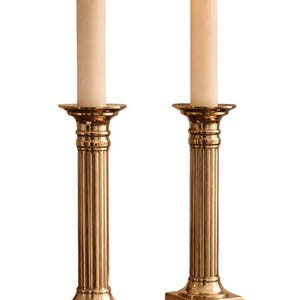 May include: Two brass candle holders with lit ivory candles. The candle holders have a fluted column design and square bases. The candles are centered on the holders, with a small flame at the top. The background is white.