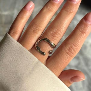 Könnte beinhalten: Ein silberfarbener Ring mit einem abstrakten, fließenden Design. Der Ring hat eine einzigartige Form mit abgerundeten Kanten und einer Lücke in der Mitte. Der Ring wird an einem Finger mit hellrosa Nagellack getragen.