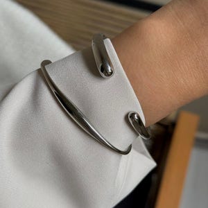 Könnte beinhalten: Ein poliertes silbernes Armreif-Armband mit minimalistischem Design. Das Armband hat einen gebogenen Stab, der sich um das Handgelenk wickelt, mit tropfenförmigen Enden. Der Schmuck wird über einem weißen Langarmhemd getragen.