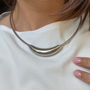 Könnte beinhalten: Eine silberfarbene Halskette mit einem gebogenen, polierten Anhänger. Die Halskette hat eine dünne, flexible Kette. Der Anhänger hat eine glatte, längliche Form und reflektiert das Licht. Die Halskette wird auf einem weißen Oberteil getragen.