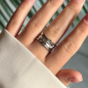 Könnte beinhalten: Ein silberner Ring mit einem wellenförmigen, organischen Design. Der Ring wird an einem Finger getragen und zeigt seine strukturierte Oberfläche. Die reflektierende Qualität des Rings fängt das Licht ein und unterstreicht seine einzigartige Form und seinen Stil.