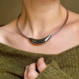 Könnte beinhalten: Eine silberfarbene Halskette mit einem gebogenen, polierten Anhänger. Die Halskette hat ein flexibles, spiralförmiges Drahtdesign. Der Anhänger hat eine glatte, abgerundete Form. Die Halskette wird von einer Person getragen.