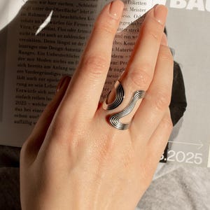 Könnte beinhalten: Ein silberfarbener Ring mit einem einzigartigen, fließenden Design wird auf einer Hand gezeigt. Der Ring hat eine Reihe von geschwungenen Linien und eine glatte, polierte Oberfläche. Das Design des Rings ist modern und auffällig, geeignet für den Alltag oder besondere Anlässe.