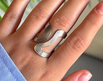 May include: A silver-colored ring with a wavy, open design. The ring features multiple parallel lines that curve and flow across the finger. The ring is worn on a hand with manicured nails, and a blue shirt sleeve is visible in the background.