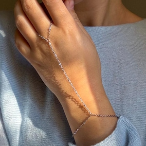Peut inclure: Un bracelet de main en argent délicat. Le bracelet est composé d'une fine chaîne qui relie le poignet au doigt, créant un design minimaliste et élégant. Le bijou est porté sur la main et le poignet.