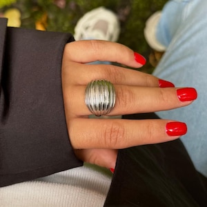 Könnte beinhalten: Ein silberfarbener Ring mit geripptem, ovalem Design. Der Ring wird an einer Hand mit rotem Nagellack getragen. Die Hand ruht auf einem schwarzen Kleidungsstück, darunter ein weißes Kleidungsstück. Der Hintergrund ist unscharf.