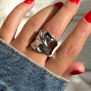 Könnte beinhalten: Ein silberfarbener Ring mit einem abstrakten, strukturierten Design. Der Ring hat eine unregelmäßige Form mit poliertem Finish. Der Hintergrund umfasst eine Jeansjacke und eine Hand mit rotem Nagellack.