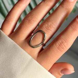 Könnte beinhalten: Ein silberfarbener Ring mit ovalem Design. Der Ring wird an einem Finger getragen, die Hand ruht auf weißem Stoff. Das Design des Rings ist schlicht und modern, mit polierter Oberfläche.