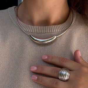 Könnte beinhalten: Eine silberfarbene Halskette mit einem gebogenen, polierten Anhänger. Die Halskette wird mit einem passenden silbernen Ring mit geripptem Design kombiniert. Der Schmuck wird vor einem neutralfarbenen Pullover präsentiert.