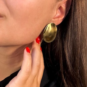 Può includere: Orecchini ovali color oro. Gli orecchini hanno una superficie leggermente strutturata e una finitura spazzolata. L'immagine mostra un orecchino a fuoco, indossato su un orecchio. La mano della persona tocca l'orecchino. La persona indossa un top nero.