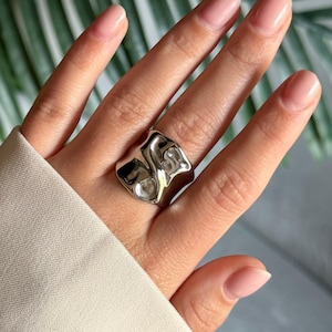 Könnte beinhalten: Ein silberfarbener Ring mit einem abstrakten, organischen Design. Der Ring hat eine strukturierte Oberfläche und wird am Finger getragen. Der Ring ist ein auffälliges Schmuckstück.