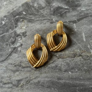 May include: A pair of gold-toned earrings with a textured, woven design. Each earring features a rectangular top connected to a rounded, heart-shaped bottom. The earrings are displayed on a gray marble surface.