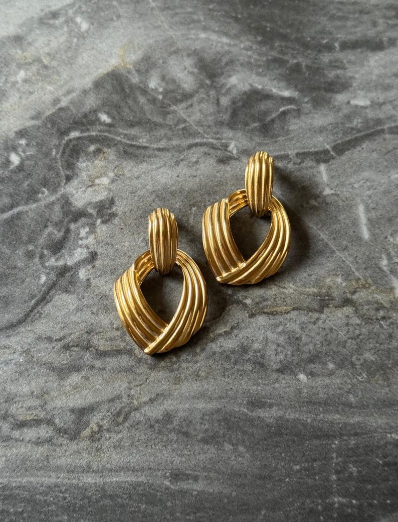 May include: A pair of gold-toned earrings with a textured, woven design. Each earring features a rectangular top connected to a rounded, heart-shaped bottom. The earrings are displayed on a gray marble surface.