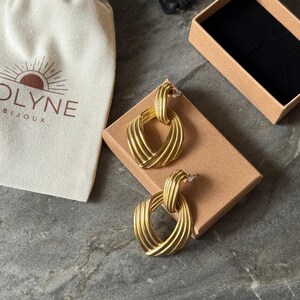 May include: A pair of gold-colored earrings with a textured, ridged design, presented on a small brown box. A cream-colored pouch with the brand name "OLYNE BIJOUX" is visible. The earrings have a post-back closure.