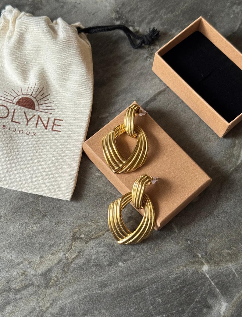 May include: A pair of gold-colored earrings with a textured, ridged design, presented on a small brown box. A cream-colored pouch with the brand name "OLYNE BIJOUX" is visible. The earrings have a post-back closure.