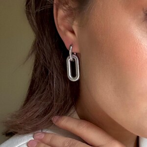 May include: Silver-toned chain link earrings. The earrings feature a small, circular top loop connected to a larger, elongated oval link. The oval link has a textured, ridged design. The earrings are shown on a person's ear.