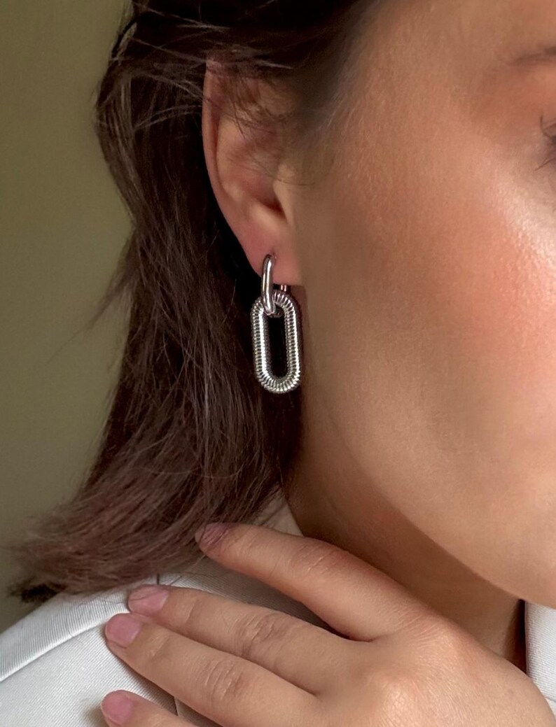 May include: Silver-toned chain link earrings. The earrings feature a small, circular top loop connected to a larger, elongated oval link. The oval link has a textured, ridged design. The earrings are shown on a person's ear.