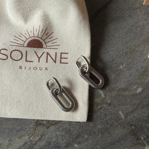 May include: Silver-toned oval hoop earrings with a textured, coiled design. The earrings are displayed next to a cream-colored pouch with the brand name "SOLYNE BIJOUX" printed on it. The earrings are on a gray surface.