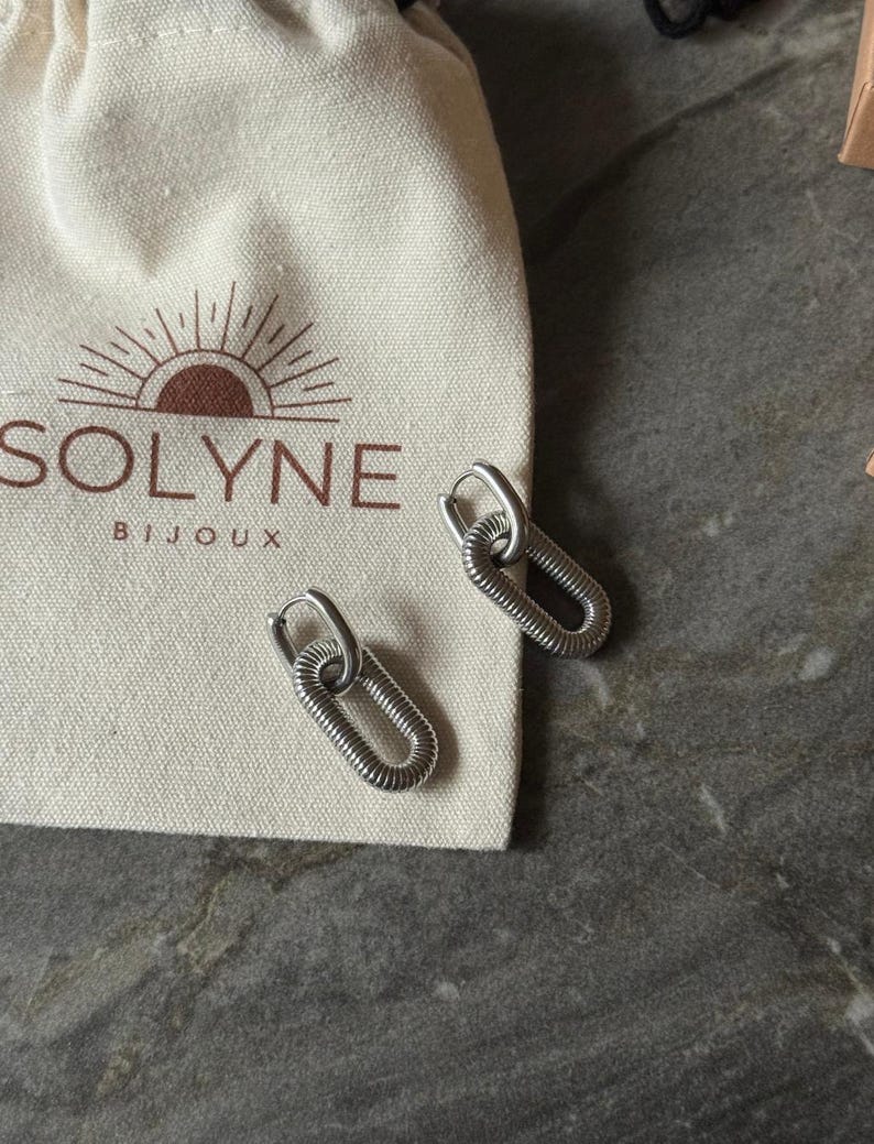 May include: Silver-toned oval hoop earrings with a textured, coiled design. The earrings are displayed next to a cream-colored pouch with the brand name "SOLYNE BIJOUX" printed on it. The earrings are on a gray surface.