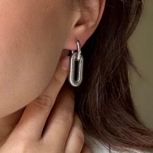 May include: Silver-toned chain link earrings. The earrings have a textured, ribbed design and a hinged closure. The oval-shaped links are connected to a smaller, circular loop that attaches to the ear. The earrings are shown on a person's ear.