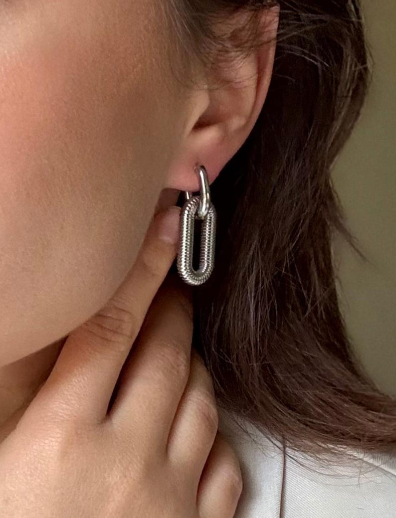 May include: Silver-toned chain link earrings. The earrings have a textured, ribbed design and a hinged closure. The oval-shaped links are connected to a smaller, circular loop that attaches to the ear. The earrings are shown on a person's ear.