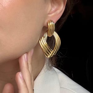 May include: Gold-toned, textured earrings with a geometric design. The earrings feature a series of curved, interwoven lines, creating a layered effect. The earrings are attached to the ear with a post. The background is dark.