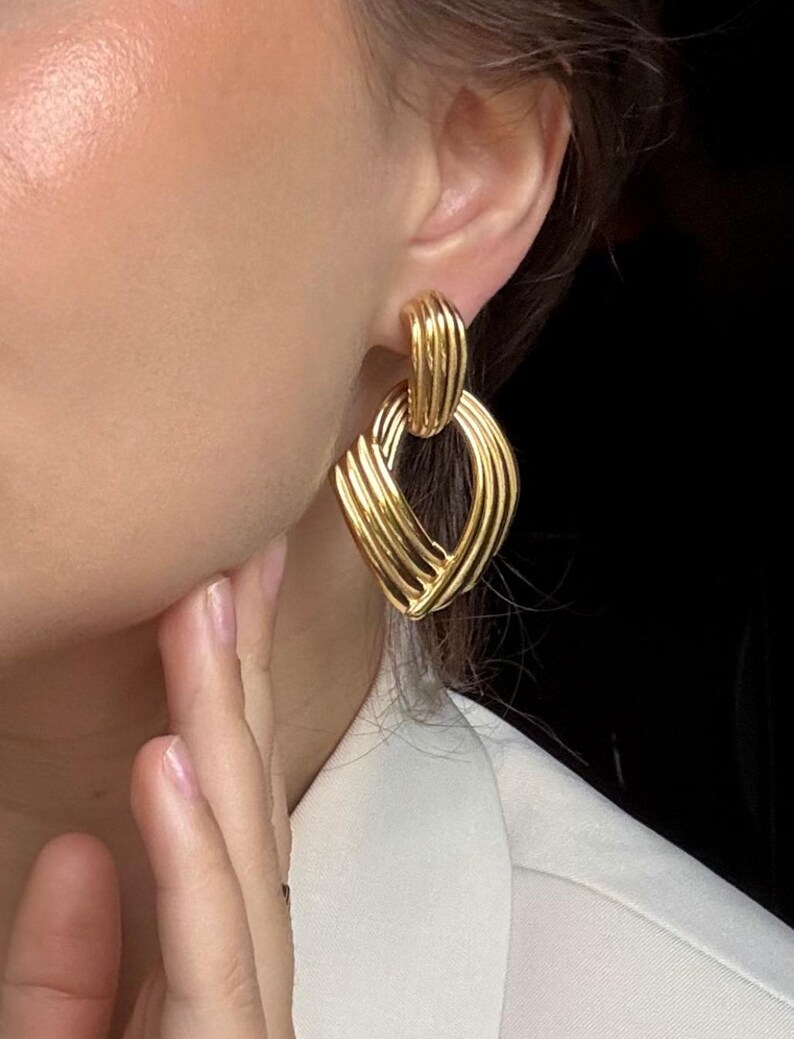 May include: Gold-toned, textured earrings with a geometric design. The earrings feature a series of curved, interwoven lines, creating a layered effect. The earrings are attached to the ear with a post. The background is dark.