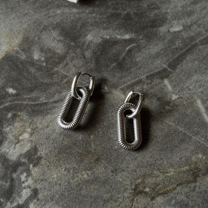 May include: A pair of silver-toned earrings. Each earring features a small hoop that connects to a larger, elongated oval shape with a textured, coiled design. The earrings are displayed on a gray, marbled surface.