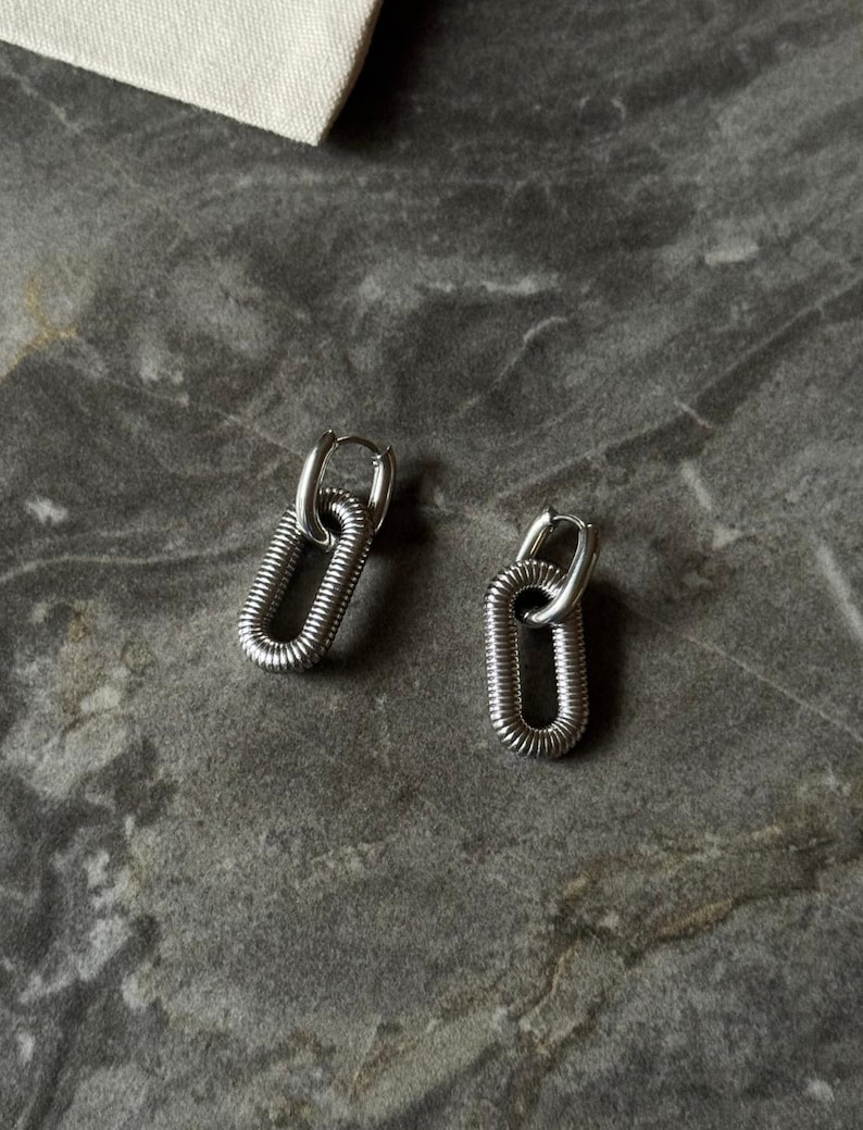 May include: A pair of silver-toned earrings. Each earring features a small hoop that connects to a larger, elongated oval shape with a textured, coiled design. The earrings are displayed on a gray, marbled surface.