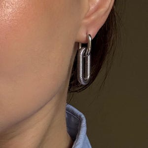 May include: Silver-toned, chain-link style earrings. The earrings feature a small hoop that connects to a larger, elongated oval link. The oval link has a textured, ridged design. The earrings are worn on the ear.
