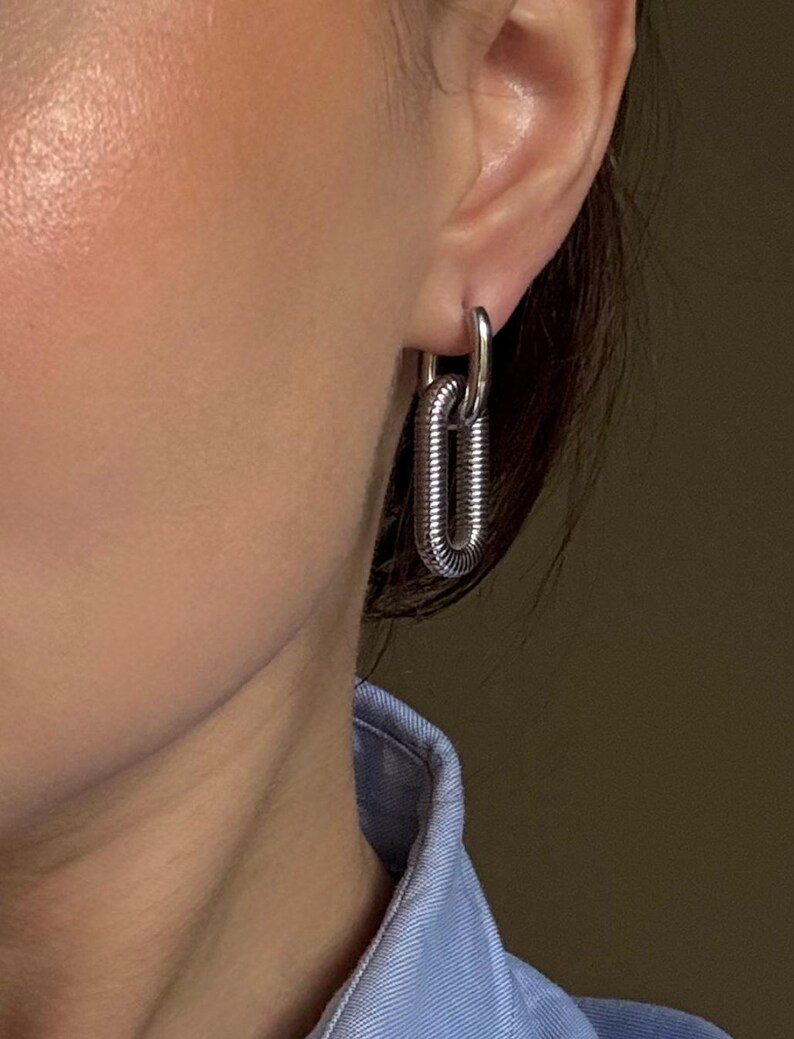 May include: Silver-toned, chain-link style earrings. The earrings feature a small hoop that connects to a larger, elongated oval link. The oval link has a textured, ridged design. The earrings are worn on the ear.