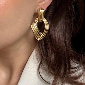 May include: Gold-toned earrings with a geometric design. The earrings feature a rectangular shape with a series of curved lines, creating a textured, woven appearance. The earrings are attached to a smaller, ribbed loop.