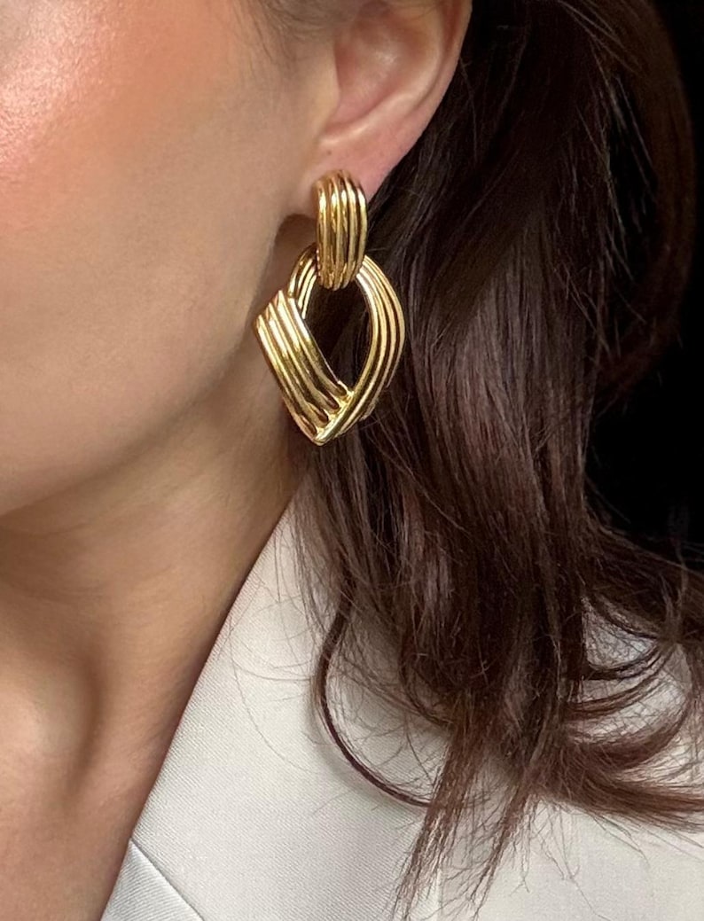 May include: Gold-toned earrings with a geometric design. The earrings feature a rectangular shape with a series of curved lines, creating a textured, woven appearance. The earrings are attached to a smaller, ribbed loop.
