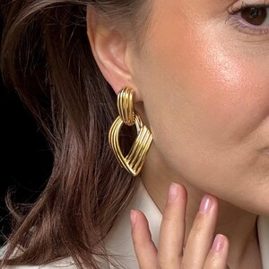 May include: Gold-toned earrings with a geometric design. The earrings feature a series of parallel lines, creating a textured, layered effect. A matching gold ring is also visible. The jewelry is worn on a person.