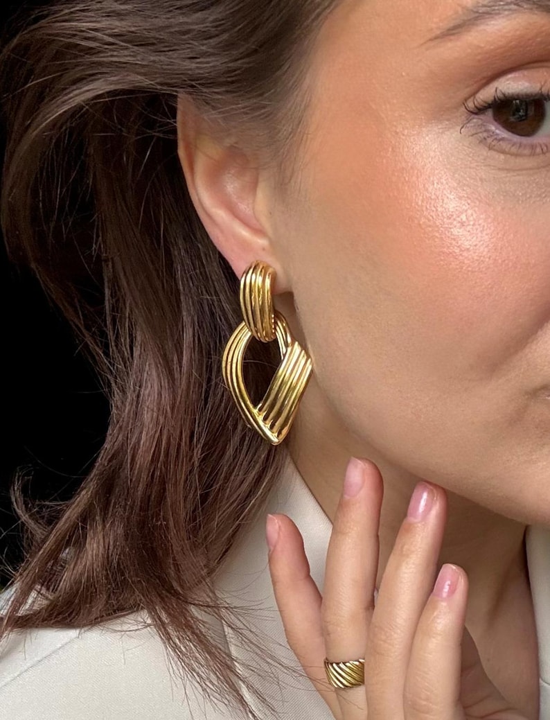 May include: Gold-toned earrings with a geometric design. The earrings feature a series of parallel lines, creating a textured, layered effect. A matching gold ring is also visible. The jewelry is worn on a person.