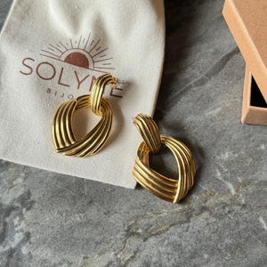 May include: A pair of gold-colored earrings with a woven design. The earrings are in the shape of a heart and are displayed on a gray surface next to a small beige bag with the brand name "SOLYME BIJO" printed on it.
