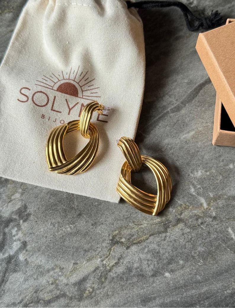 May include: A pair of gold-colored earrings with a woven design. The earrings are in the shape of a heart and are displayed on a gray surface next to a small beige bag with the brand name "SOLYME BIJO" printed on it.