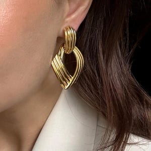 May include: Gold-toned earrings with a geometric design. The earrings feature a ribbed, oval-shaped top that connects to a diamond-shaped pendant with a layered, textured appearance. The earrings are a bright, shiny gold color.