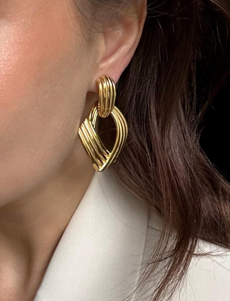 May include: Gold-toned earrings with a geometric design. The earrings feature a ribbed, oval-shaped top that connects to a diamond-shaped pendant with a layered, textured appearance. The earrings are a bright, shiny gold color.