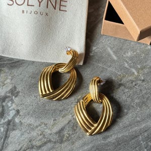 May include: A pair of gold-colored earrings with a textured, woven design. The earrings have a post-back closure and a geometric shape. The earrings are displayed on a gray surface next to a white bag with the text "SOLYNE BIJOUX" and a brown box.