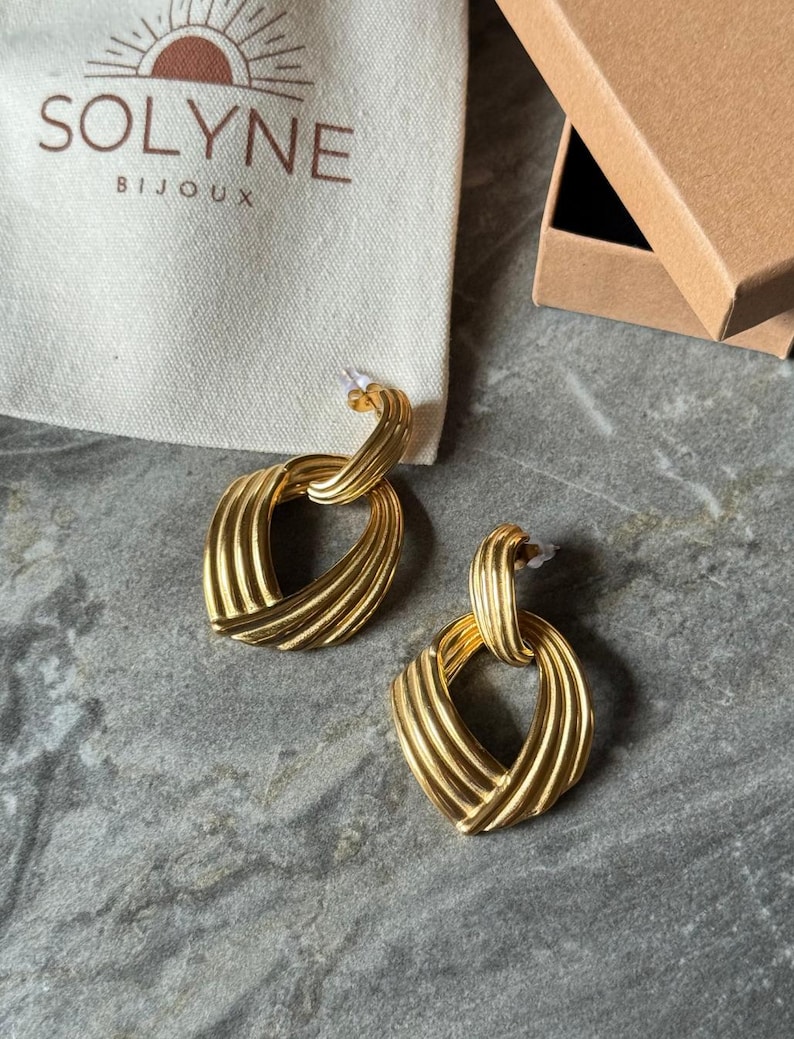 May include: A pair of gold-colored earrings with a textured, woven design. The earrings have a post-back closure and a geometric shape. The earrings are displayed on a gray surface next to a white bag with the text "SOLYNE BIJOUX" and a brown box.