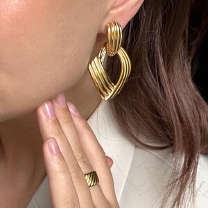 May include: Gold-toned earrings and ring set. The earrings feature a geometric design with a textured, ribbed pattern. The ring has a matching design. The jewelry is worn on a person.