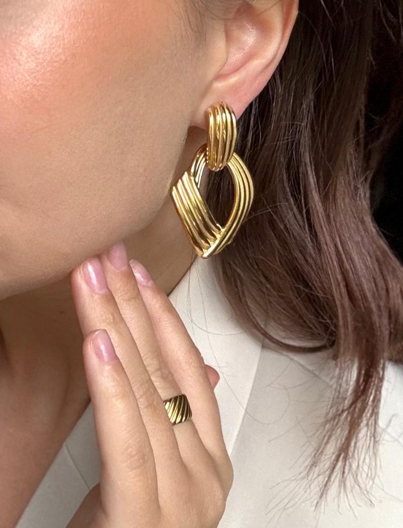 May include: Gold-toned earrings and ring set. The earrings feature a geometric design with a textured, ribbed pattern. The ring has a matching design. The jewelry is worn on a person.