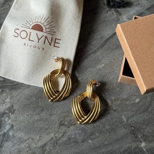 May include: Gold-toned earrings with a woven heart design, displayed with a drawstring pouch and a small cardboard box. The pouch has the text "SOLYNE BIJOUX" and a sun logo. The earrings are on a gray surface.
