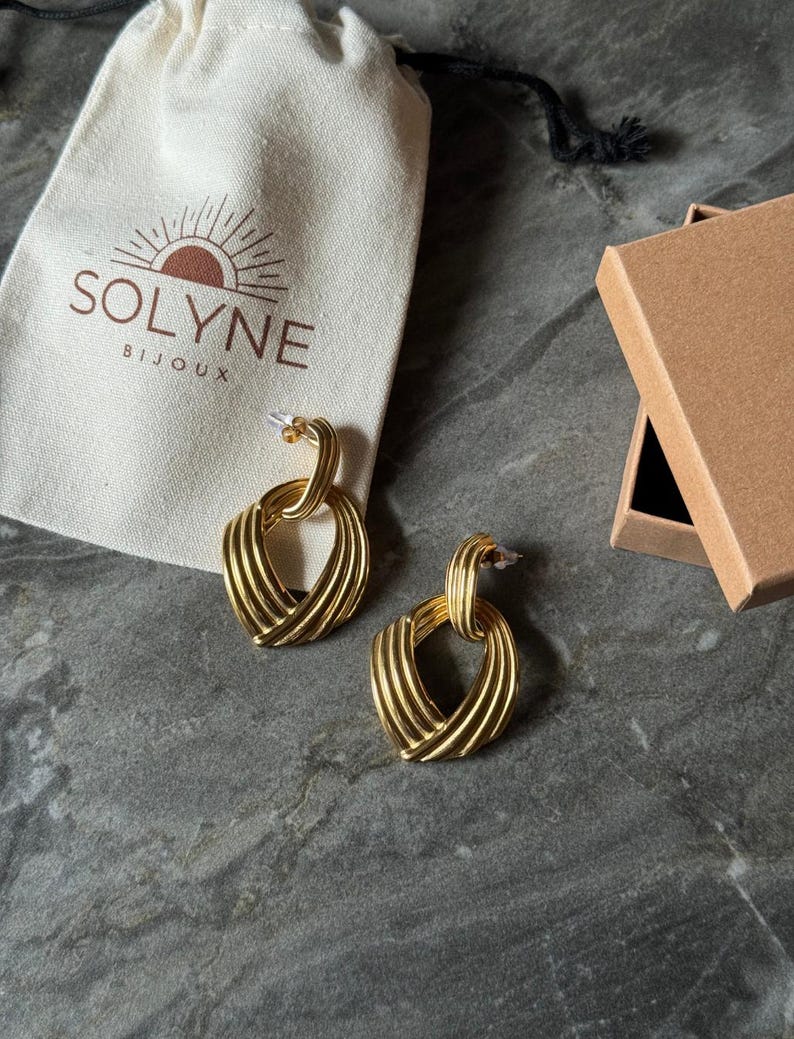 May include: Gold-toned earrings with a woven heart design, displayed with a drawstring pouch and a small cardboard box. The pouch has the text "SOLYNE BIJOUX" and a sun logo. The earrings are on a gray surface.