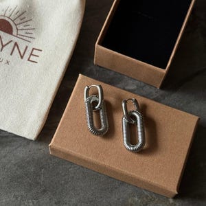 May include: A pair of silver-toned, oval-shaped earrings with a textured, coiled design. The earrings are displayed on a brown cardboard box, next to a small canvas bag with a sun logo and the word "YNE".