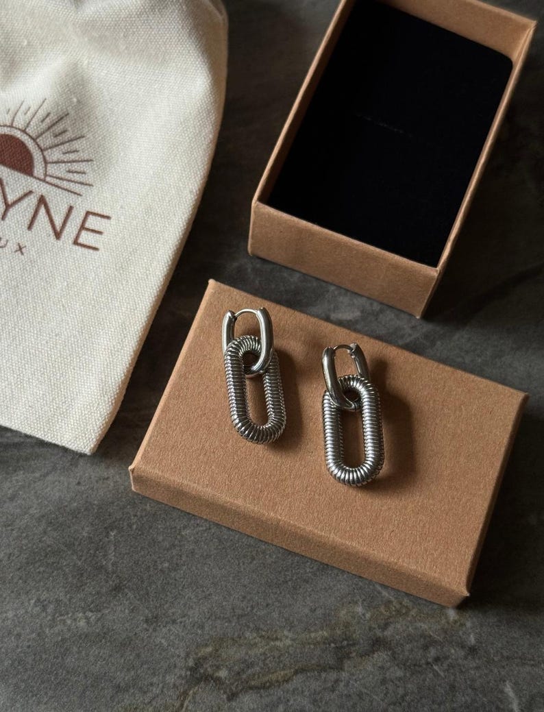 May include: A pair of silver-toned, oval-shaped earrings with a textured, coiled design. The earrings are displayed on a brown cardboard box, next to a small canvas bag with a sun logo and the word "YNE".